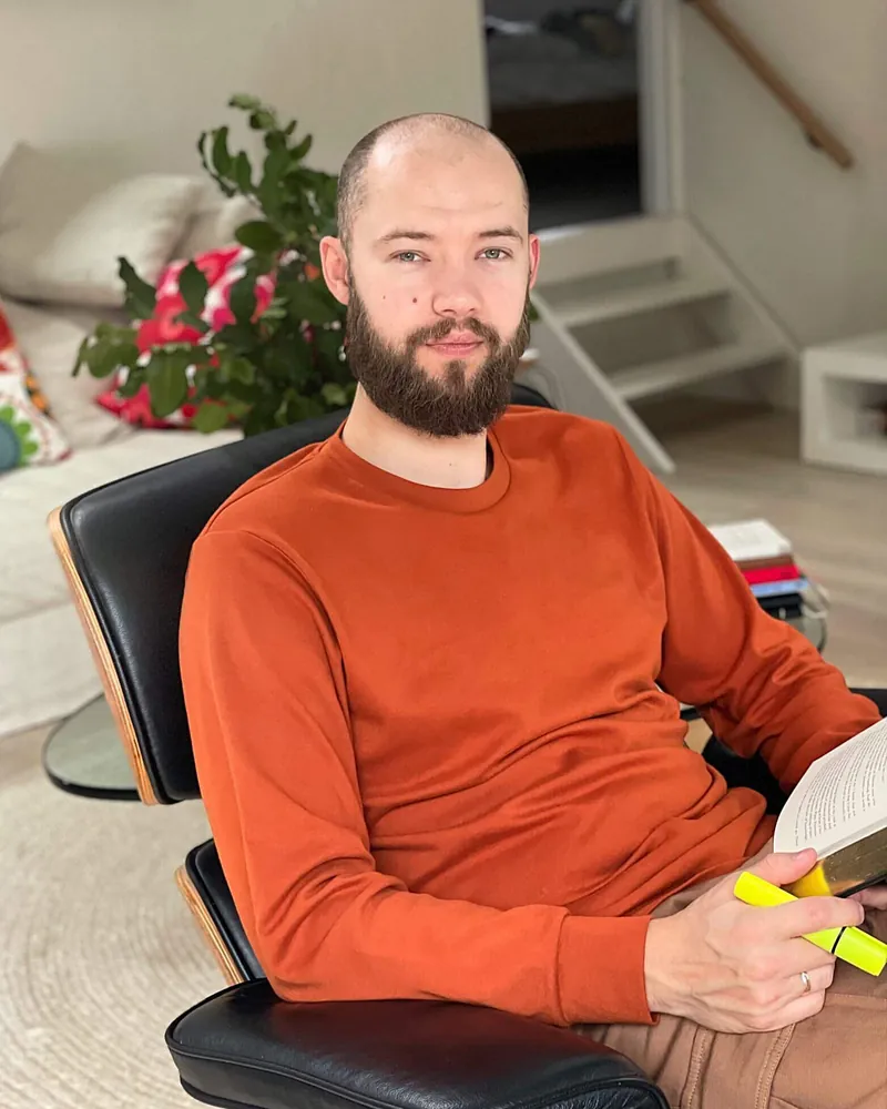 Vlad Kutepov in an orange sweater, sitting in a leather lounge chair with a book